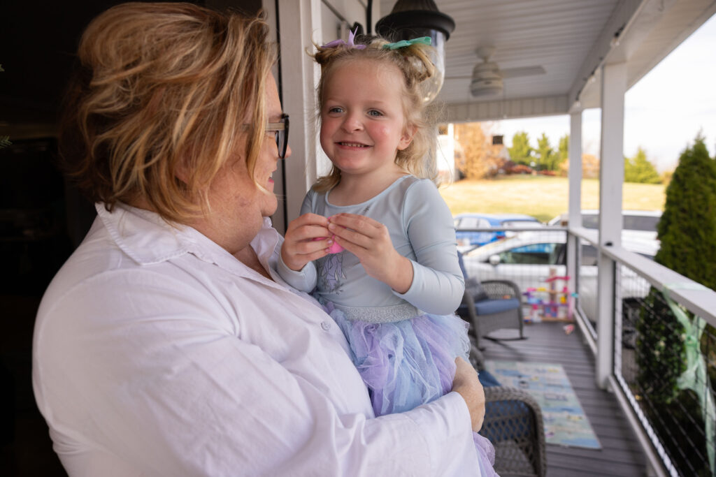 A mother holds her 3-year-old daughter in her arms on their porch. The daughter is wearing a big smile.