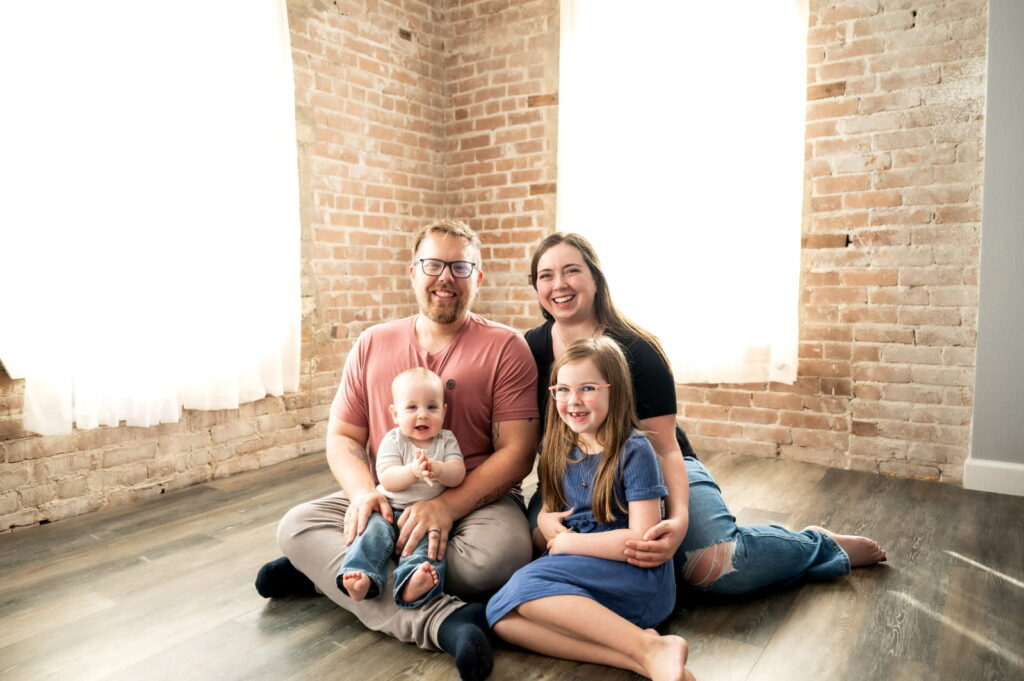 A mother and father sit on the floor in a well-lit room with their two young children in their laps for a studio portrait.