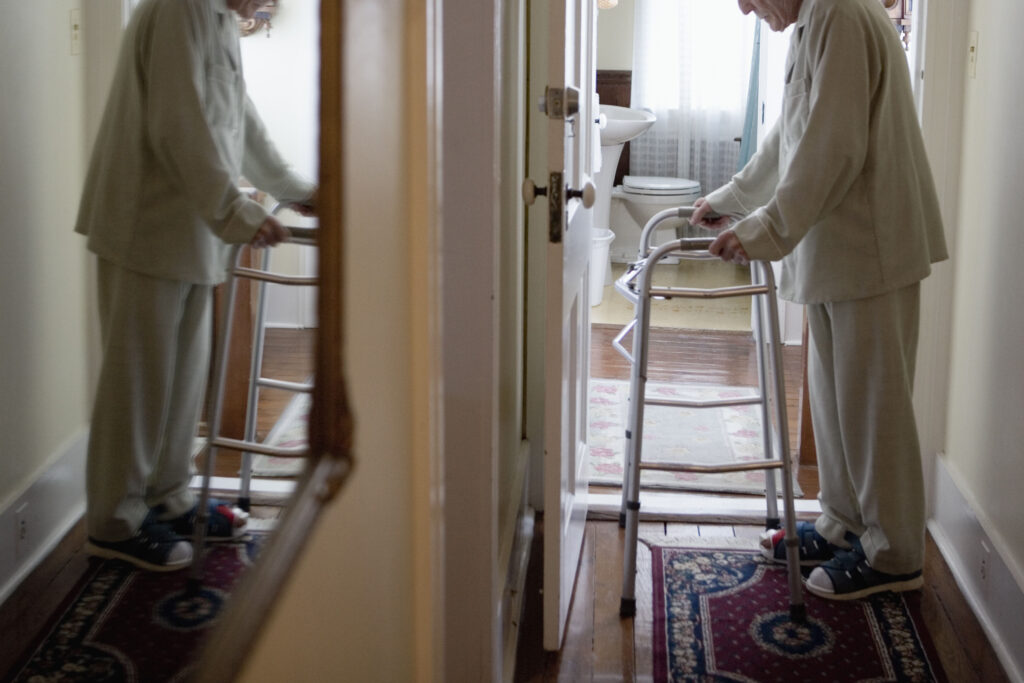 A senior man stands in a hallway with a walker. His reflection is visible, slightly blurred, in a mirror across from him.