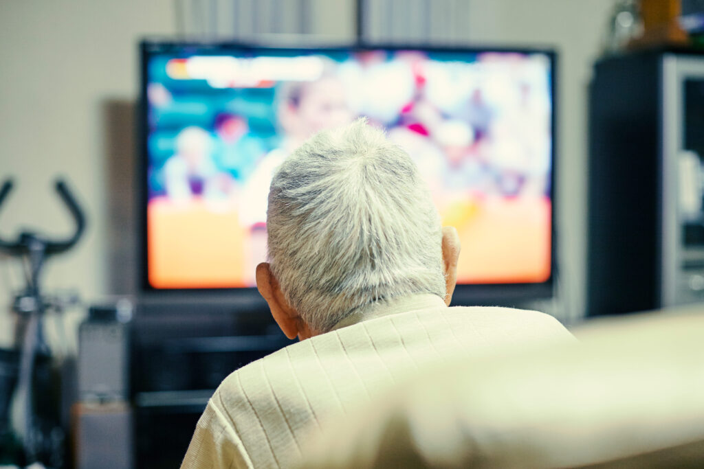 An older man is seen from behind lounging in a chair. A TV is on in front of him.