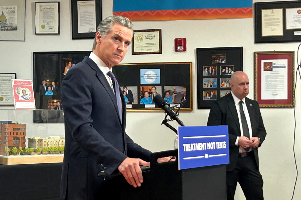 Gavin Newsom speaks at a news conference behind a podium with a sign on it that reads, "Treatment not tents."
