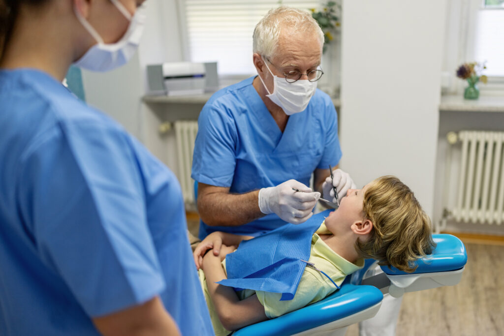 An older male dentist performs a cleaning on a young boy patient. A female hygienist is seen blurred in the foreground.