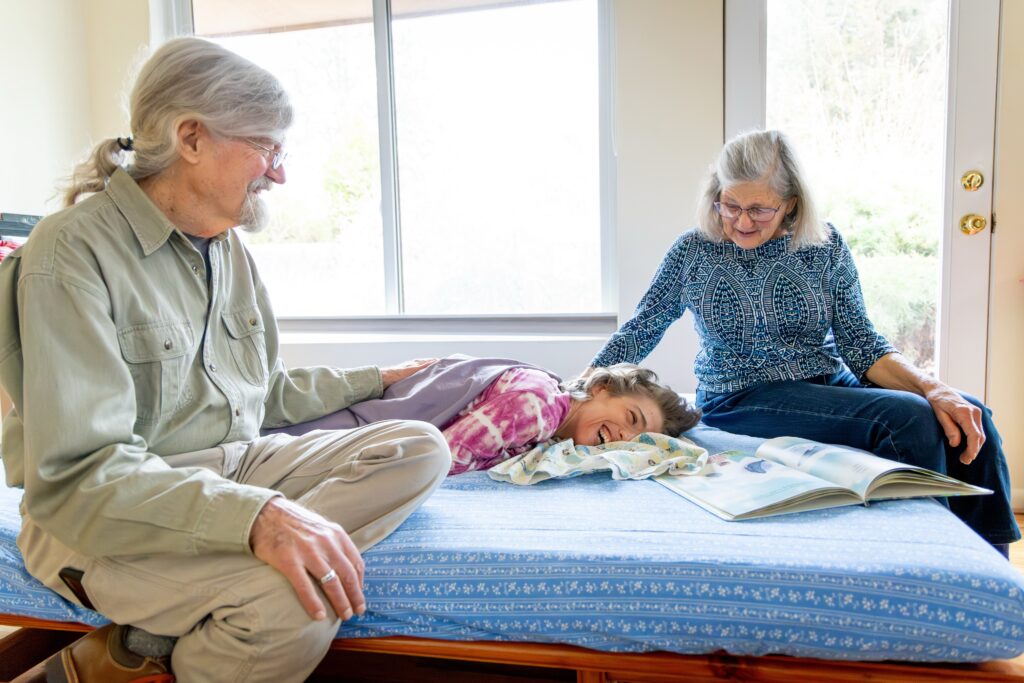 An older mother and father sit on a bed beside a smiling woman, laying down. The woman is living with a disability.