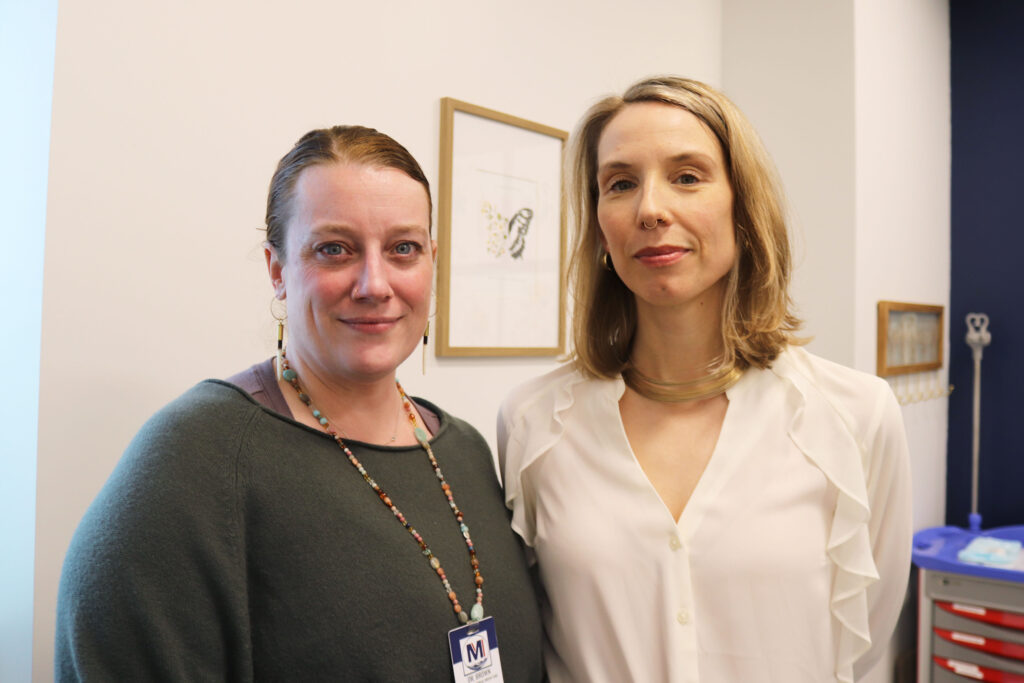 Two women with blonde-ish hair stand side-by-side in a doctor's office.